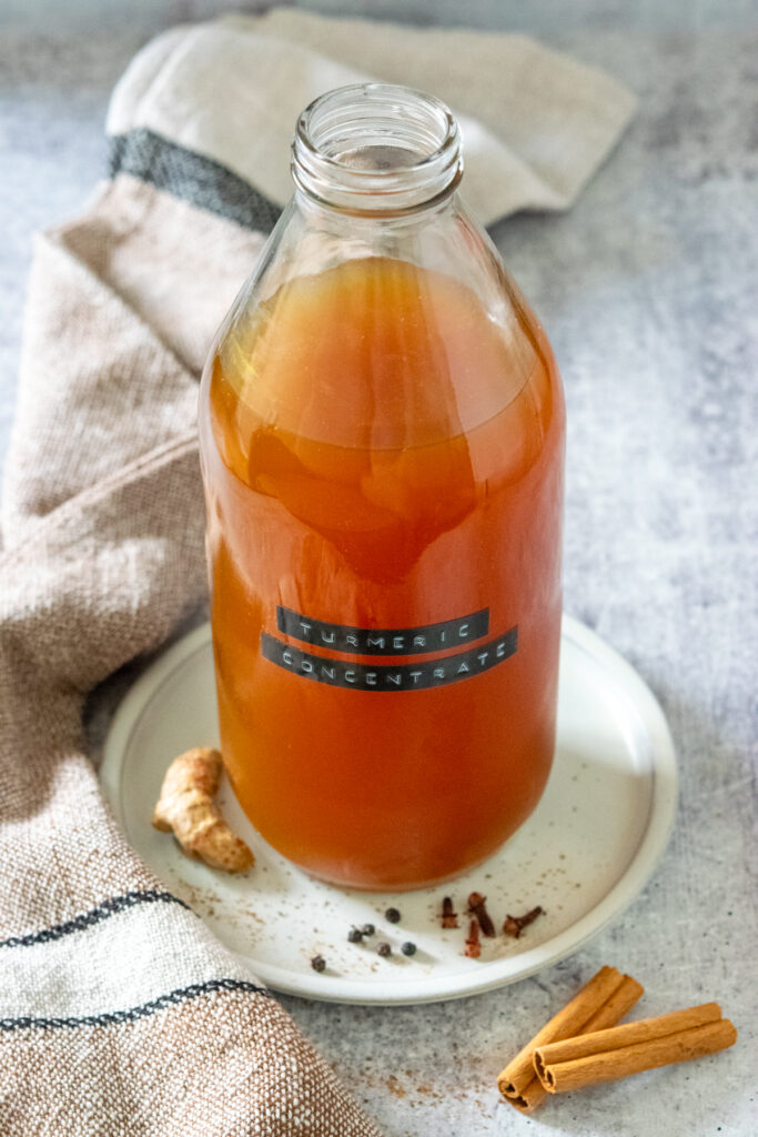 A glass bottle filled with 32 ounces of turmeric tea concentrate. The bottle is on a small plate that has turmeric root, some black peppercorns, cloves and cinnamon sticks on it.