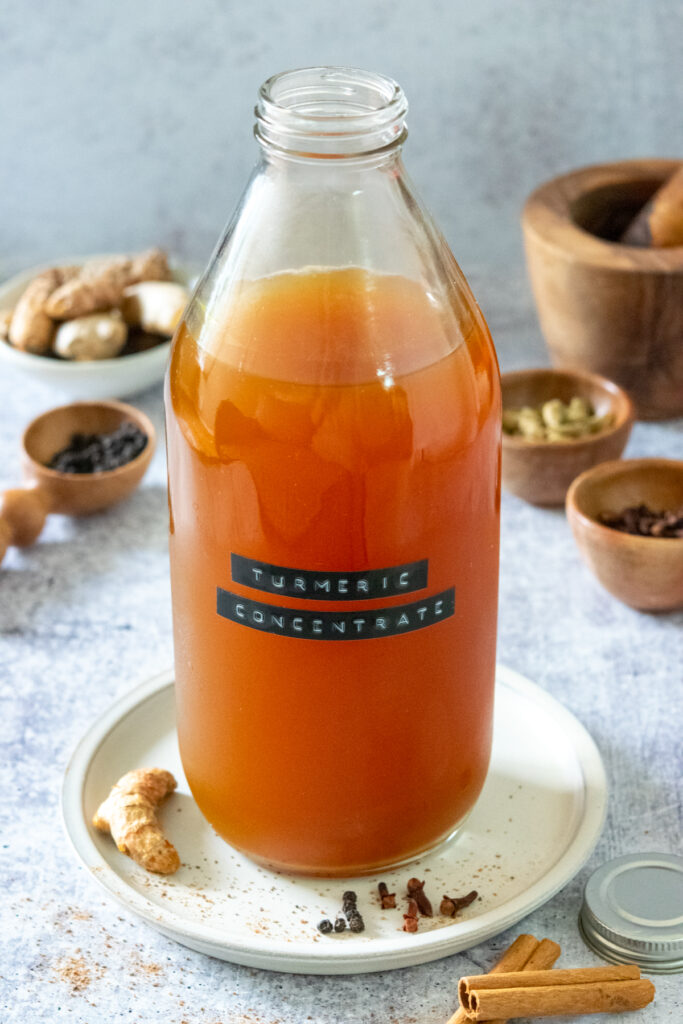 A bowl filled with fresh turmeric next to a bottle of homemade turmeric concentrate for making tea drinks. There is also some ginger root pictured and some small bowls filled with whole spices like black peppercorn, cardamom and cloves.