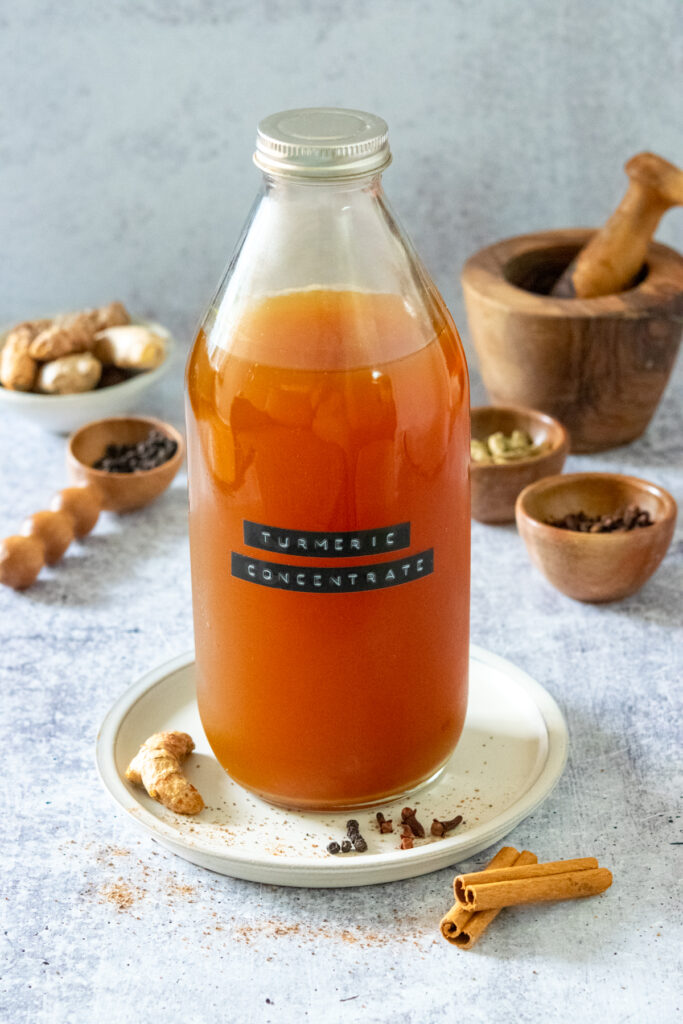 A bottle of homemade turmeric tea concentrate. The bottle is labeled "turmeric concentrate." There is a finger of turmeric root next to the tea and there is ginger and whole spices like cinnamon and cloves behind the bottle.