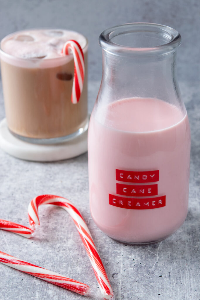 A bottle of homemade candy cane creamer next to a coffee drink topped with pink candy cane cold foam and garnished with a candy cane.