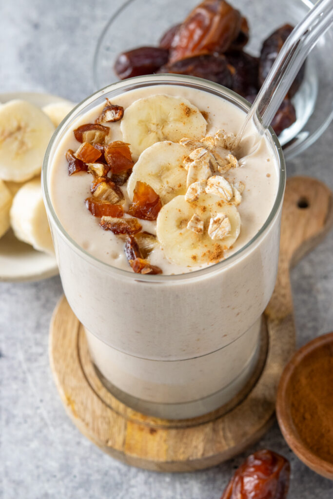A banana date breakfast smoothie in a glass with a straw next to a bowl of frozen bananas and a bowl of medjool dates. The thick smoothie is garnished with dates, bananas, oats and cinnamon.