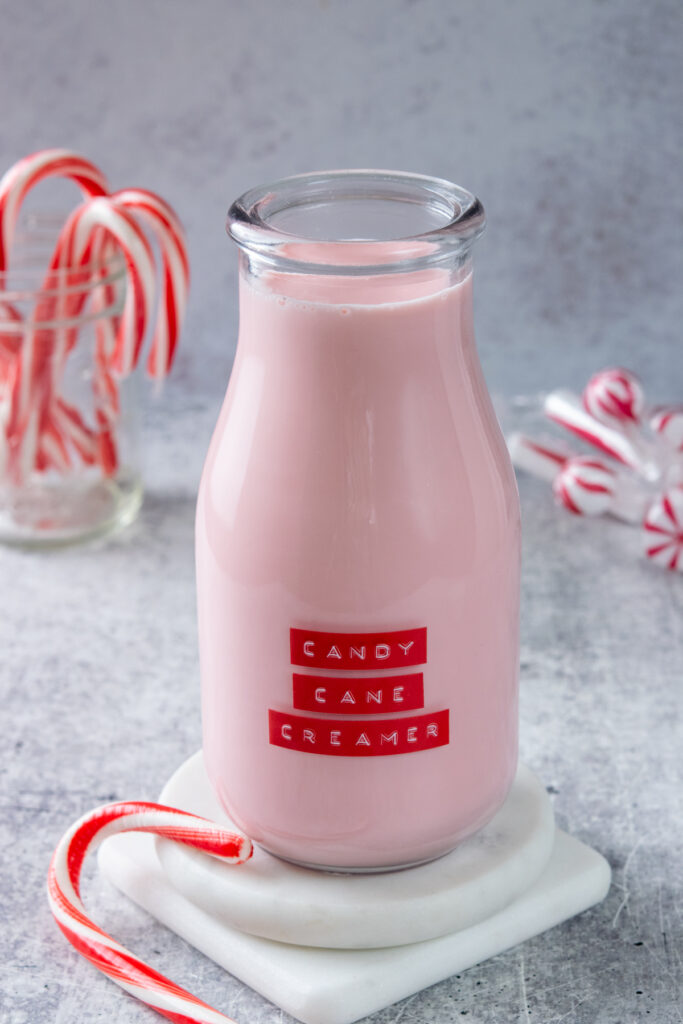 A bottle of homemade candy cane coffee creamer that is pink and has a red label on it. There is a candy cane next to the creamer and a jar filled with candy canes behind it.