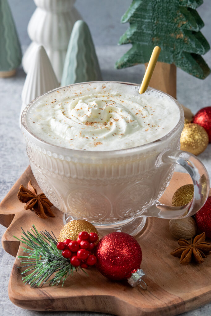 A beautiful Christmas morning chai in a embellished glass mug with a gold spoon in it. The creamy hot drink which is light tan on the bottom with a white layer of whipped cream and nutmeg, is on a Christmas tree-shaped wooden tray that has red and gold ornaments and a pine needle decoration with red berries. There's also a whole nutmeg and a couple star anise. Behind the drink are some small Christmas tree decorations showing how perfect this cozy drink is for the holidays.