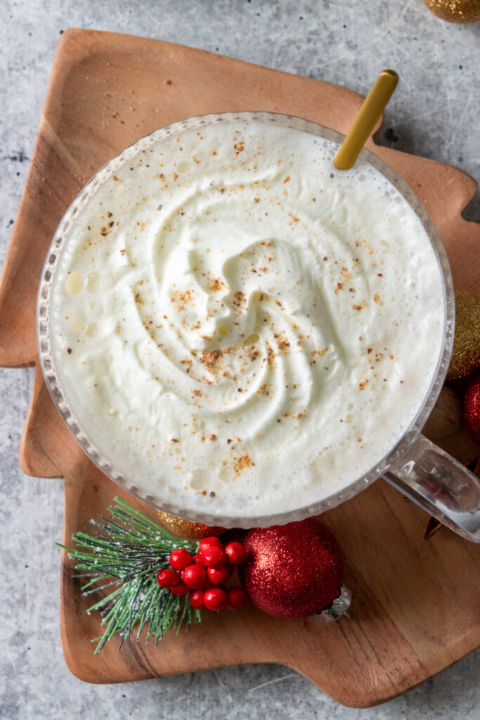 An overhead photo of the top of a Christmas morning chai. The dollop of whipped cream has melted some into a soft top like Dutch Bros, but you can still see swirls and bits of fresh nutmeg sprinkled on top. The drink is on a wood Christmas tree-shaped tray that has a few red and green Christmas ornaments on it. The drink looks very festive.