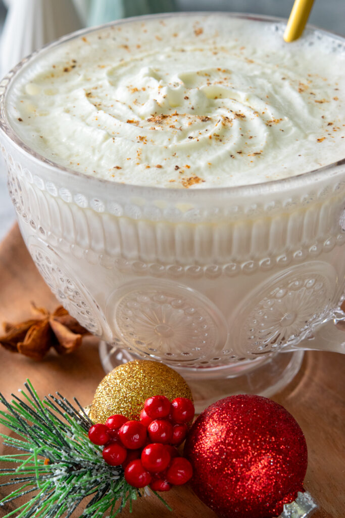 A creamy and decadent Christmas morning chai with whipped cream and nutmeg in an embellished glass mug. There are red and gold Christmas ornaments, a small pine branch with red berries and a star anise on the wood tray that the festive chai latte is sitting on.