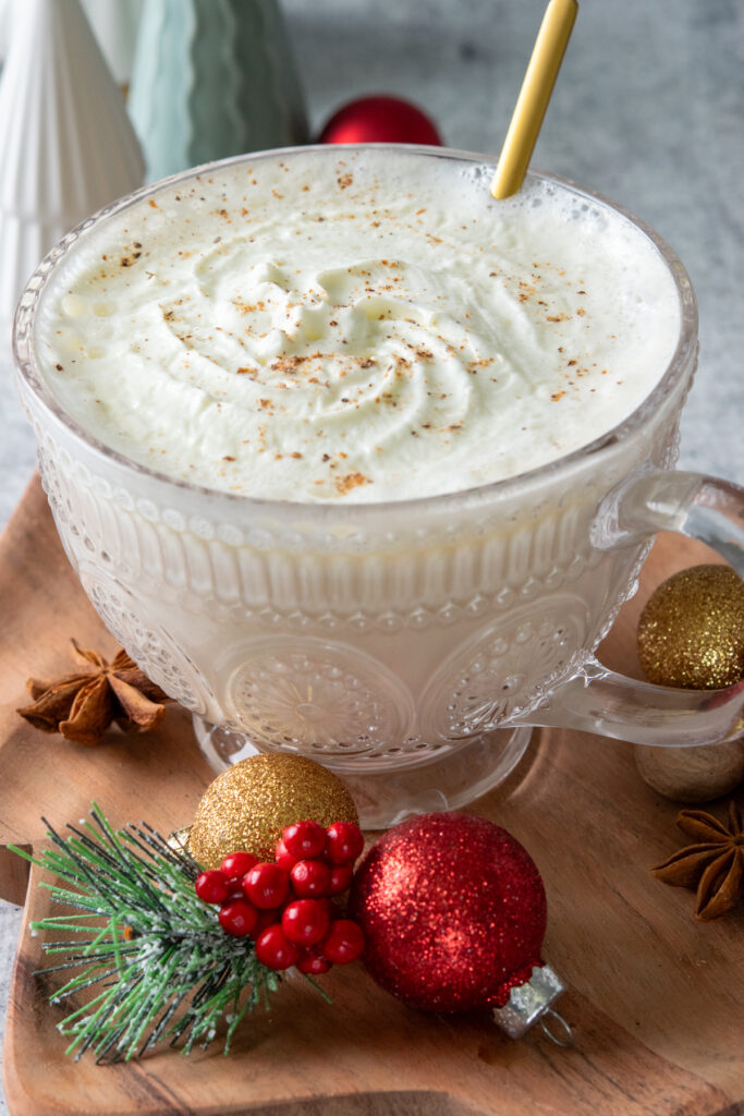 A layered warm and cozy Christmas morning chai in a glass mug with chai, white chocolate sauce and half and half on the bottom, and a thick top of semi-melted whip cream with nutmeg. There are some Christmas tree decorations and ornaments around the drink.