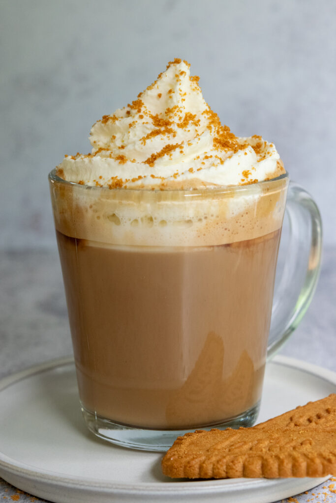 A decadent Biscoff Latte made at home showing topped with a big dollop of whipped cream and cookie crumbs, on a plate with two Lotus Biscoff cookies.
