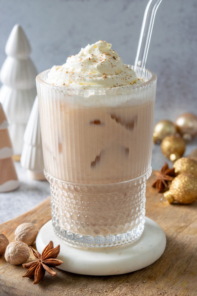 An iced chai eggnog latte in a decorative glass, topped with whipped cream and nutmeg. The holiday drink is surrounded by nutmeg, star anise, and Christmas ball and tree decorations.