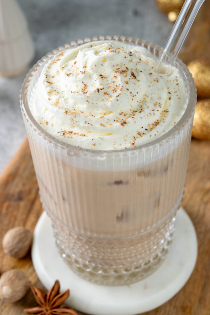 A rich and creamy eggnog chai latte topped with a thick layer of whipped cream which has freshly grated nutmeg sprinkled on top. Next to the chai tea latte are some nutmegs, star anise, and red and gold Christmas ornaments.