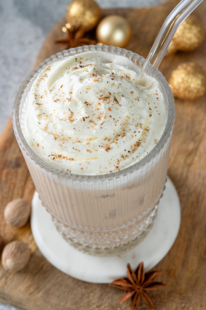 Looking down at the top of this creamy iced eggnog chai. You can see ripples of whipped cream with freshly grated nutmeg on top, The layer of cream is on top of the creamy tan-colored chai latte. Next to the drink are some whole nutmegs, a star anise and some gold Christmas balls.