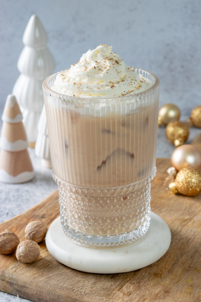 An iced eggnog chai tea latte topped with whipped cream and freshly grated nutmeg. There are some whole nutmegs near the glass and some Christmas ornaments and holiday trees behind the latte.