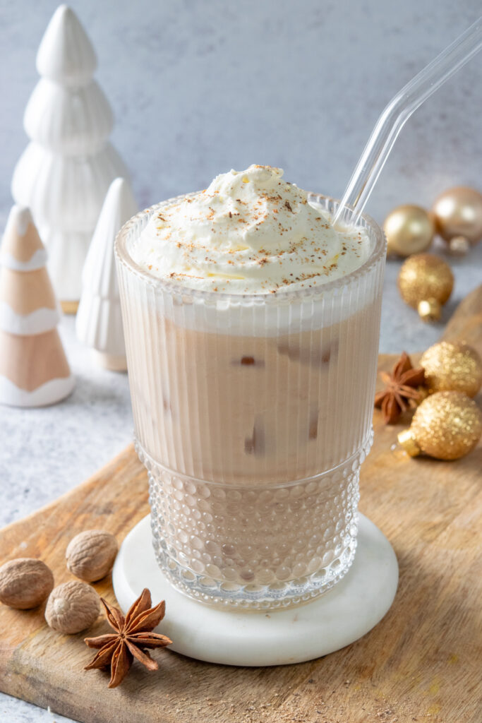 An iced eggnog chai tea latte topped with whipped cream and freshly grated nutmeg. There are some whole nutmegs near the glass and some Christmas ornaments and holiday trees behind the latte.