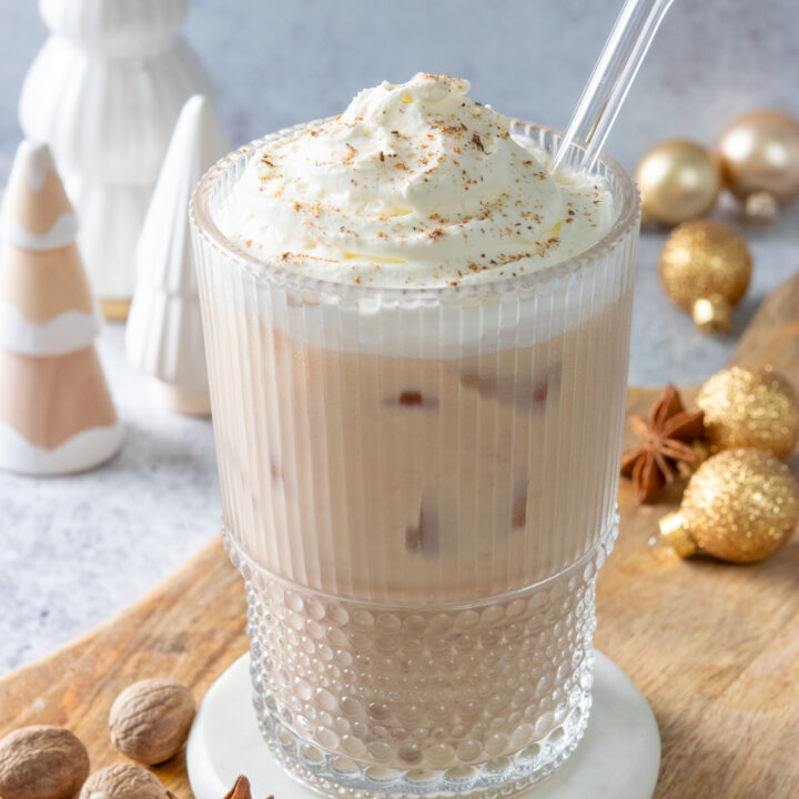 An iced eggnog chai tea latte topped with whipped cream and freshly grated nutmeg. There are some whole nutmegs near the glass and some Christmas ornaments and holiday trees behind the latte.