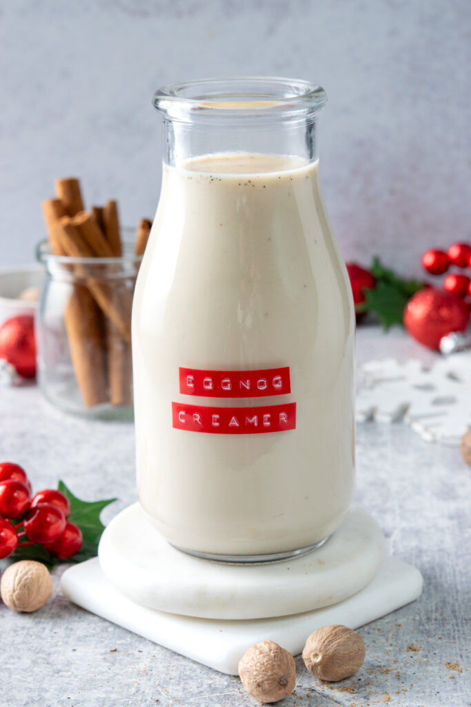 Homemade eggnog coffee creamer in a bottle. The bottle has a red label that reads, "eggnog creamer" and you can see flecks of ground nutmeg in the creamer. In front of the creamer are some whole nutmegs and behind the it is a jar with cinnamon sticks. There are some Christmas decorations strewn about.