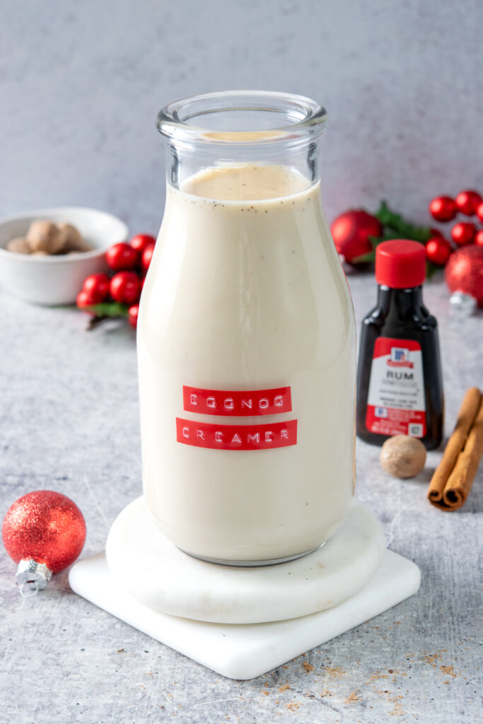 A bottle of homemade eggnog coffee creamer with a red label that reads, "eggnog creamer" Next to the creamer is a bottle of rum extract, a nutmeg, cinnamon stick and some small red Christmas ball decorations.