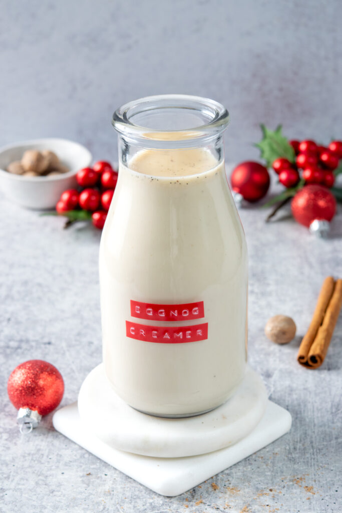 Homemade eggnog coffee creamer in a jar with a red label that reads, "eggnog creamer." Next to the creamer is a red ball Christmas ornament, a nutmeg and a cinnamon stick. Behind the creamer is a small bowl with whole nutmegs, some more red balls and a Christmas holly decoration.