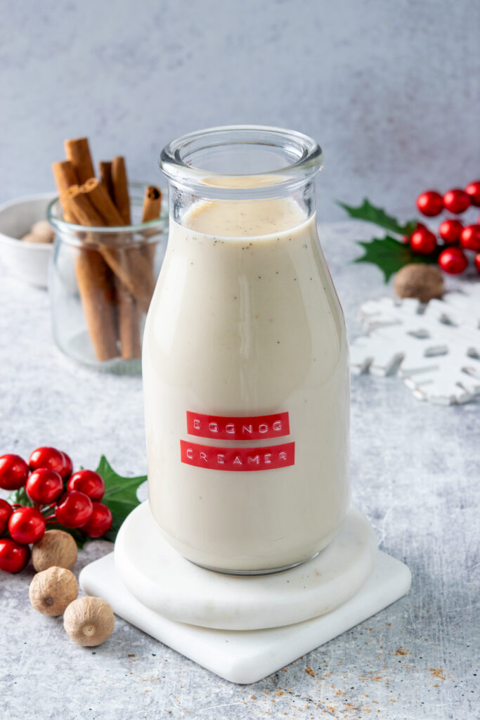 Homemade eggnog flavored creamer in a bottle that's on top of a stack of white marble coasters. Next the the bottle that has a red label are three nutmegs and a holly decoration. Behind the creamer is a jar of cinnamon sticks and a snowflake decoration and another holly.