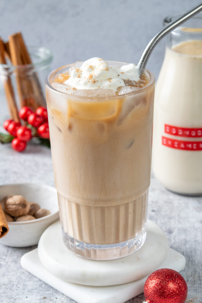 An iced eggnog latte made with the homemade creamer that's topped with a spoonful of whipped cream and freshly ground nutmeg. Behind the drink is the bottle of homemade eggnog creamer, a bowl of nutmegs and jar of cinnamon sticks. There are a few Christmas decorations scattered around the drink.