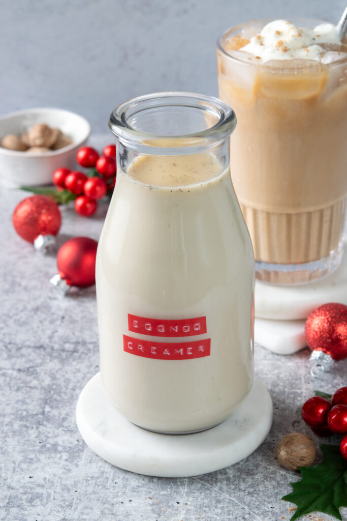 A bottle of homemade eggnog coffee creamer with a red label in front of an iced eggnog latte, some red Christmas decorations and a small bowl filled with whole nutmeg.