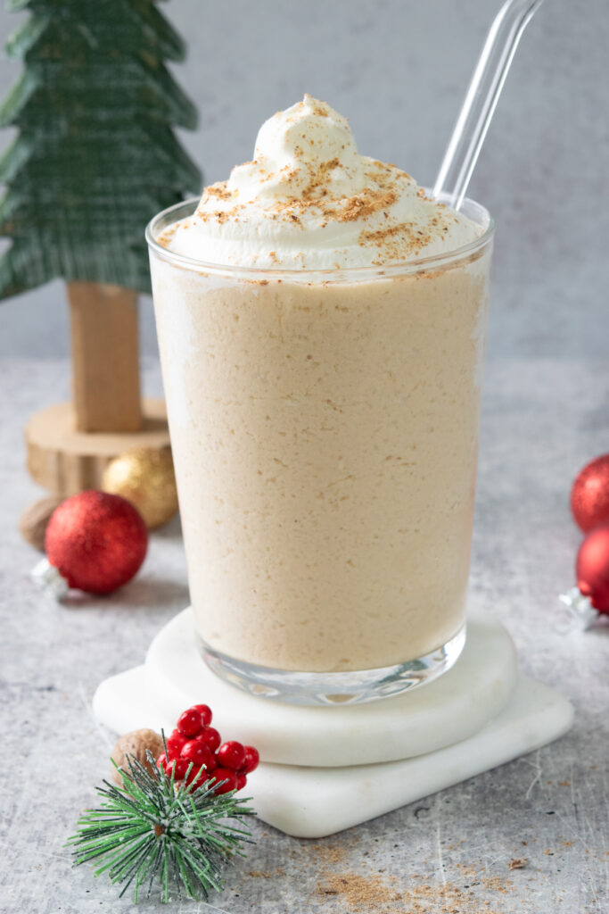 An at-home eggnog frappuccino in a glass with a glass straw. Around the drink are some small Christmas decorations including a sprig of pine needles with red berries, a Christmas tree and some red and gold ball ornaments.