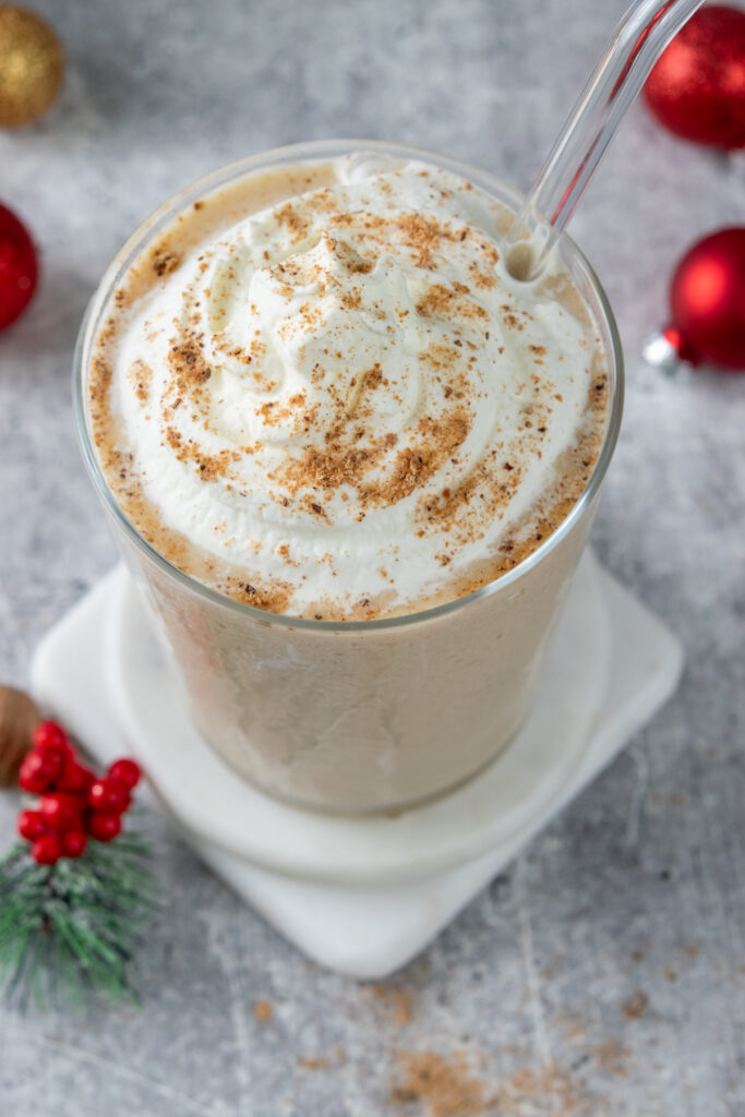 Looking at an eggnog frappuccino from the top down. The holiday frappuccino is in a glass and has a glass straw inserted into it. You can see lots of freshly ground nutmeg on the whipped cream, and the eggnog and coffee color of the thick and creamy frappuccino. The frappuccino is sitting on a stack of two white marble coasters and is surround by red, gold and green Christmas ornaments.