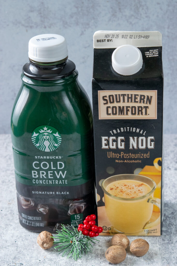 A bottle of Starbucks cold brew concentrate, which is concentrated black coffee, and a carton of non-alcoholic, traditional eggnog. In front of the two drink containers are four nutmegs and a small pine needle with red berries ornament.