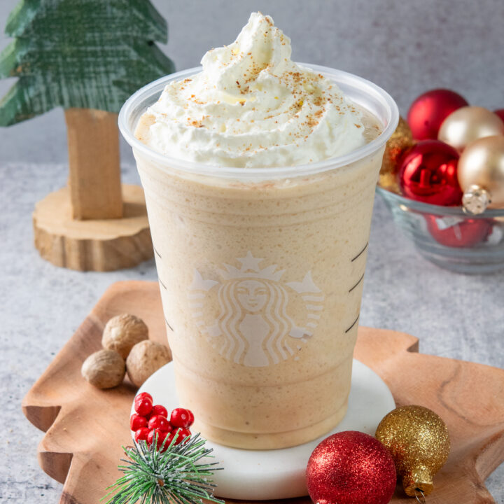 An eggnog frappuccino made at home in a Starbucks cup. The holiday drink is on a Christmas tree-shaped tray and is surrounded by festive red, green and gold ornaments.