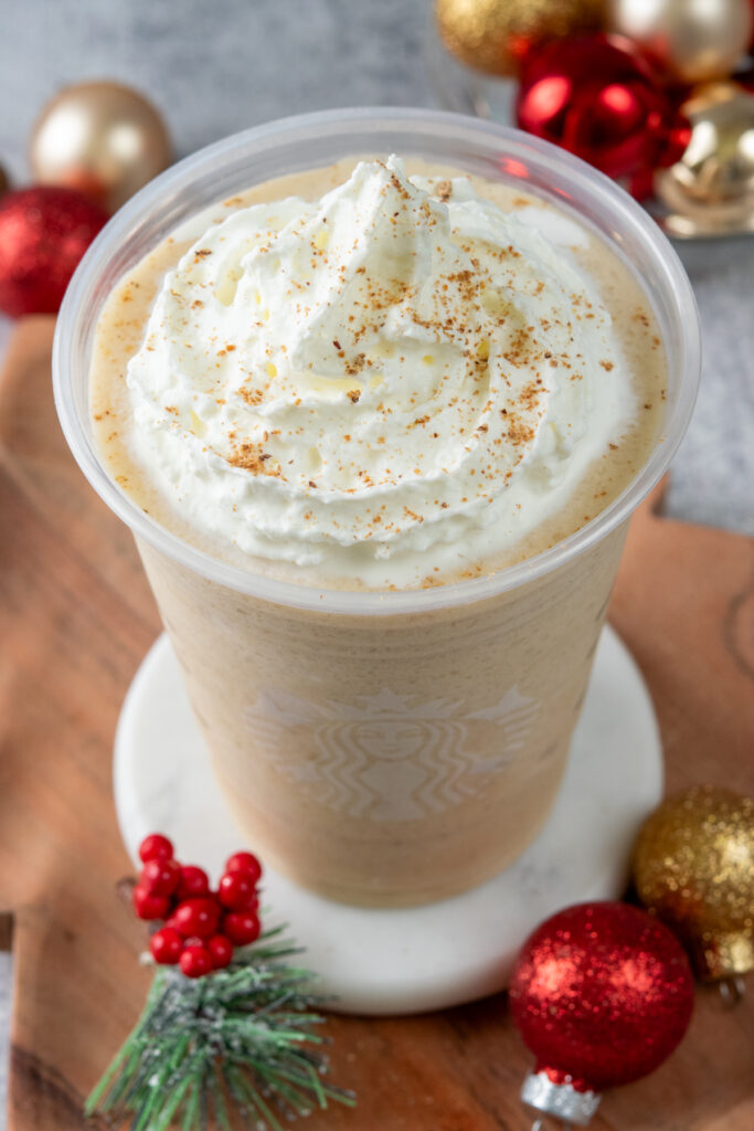 A homemade eggnog frappuccino in a Starbucks cup. You can see the top of the drink with nutmeg and whipped cream layered on top of the thick, eggnog and coffee colored frappuccino. Around the holiday drink are some small red and gold ornaments.