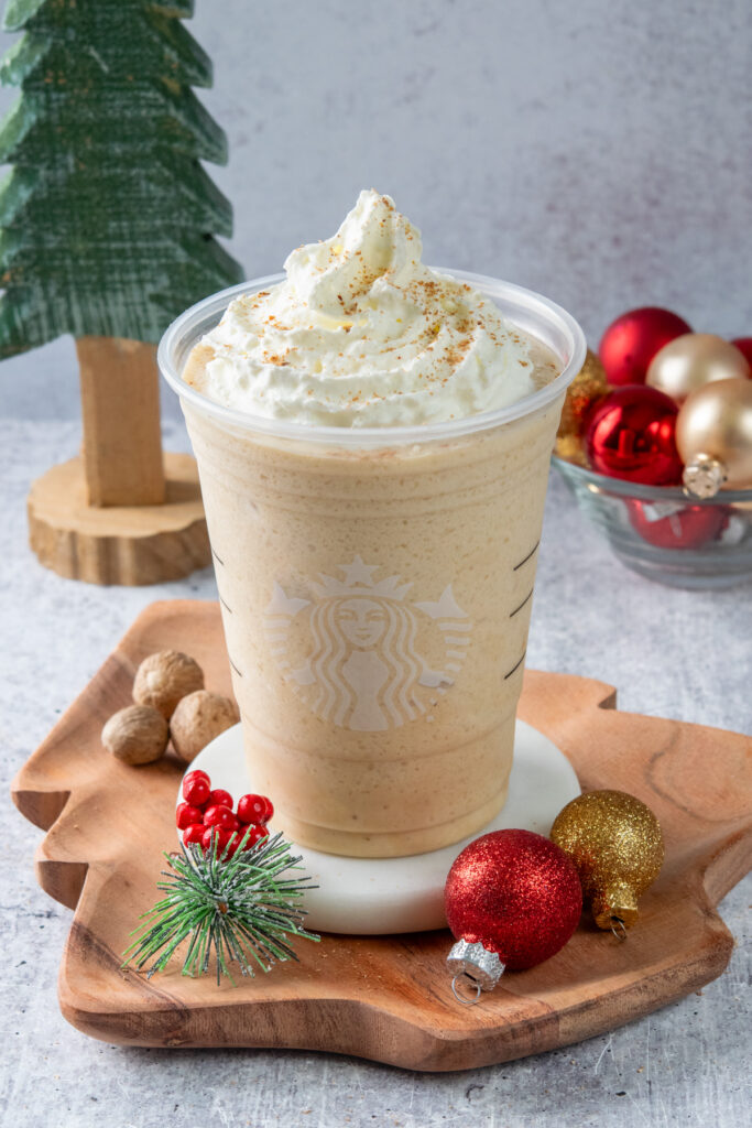 A thick and creamy eggnog frappuccino made at home in a Starbucks cup. The festive frappuccino is topped with a big swirl of whipped cream which has nutmeg sprinkled on top. Behind the copycat drink are some Christmas decorations including a Christmas tree and some small red and gold ball ornaments.