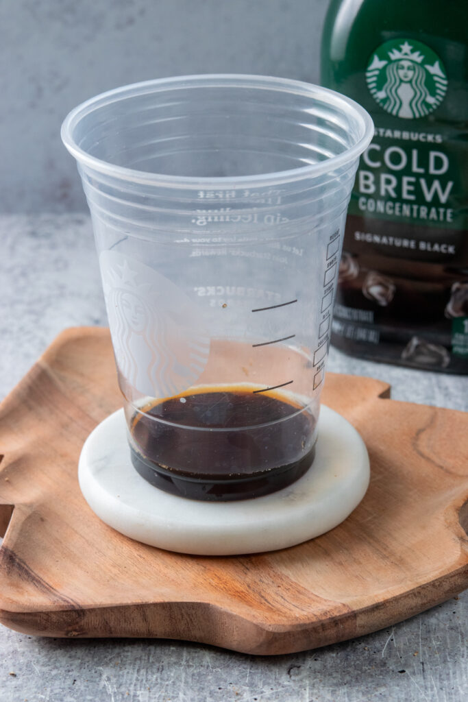 A Starbucks cold cup filled with cold brew coffee concentrate up to the first bump in the cup. There's a bottle of Starbucks cold brew concentrate behind it.