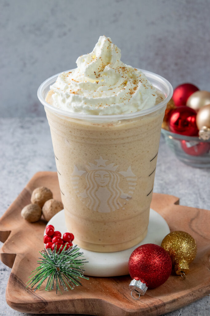 A close-up view of a homemade Starbucks eggnog frappuccino in a Starbucks cup. The festive frappuccino is sitting on a wooden Christmas-tree shaped tray which has some whole nutmegs and small red and gold Christmas ball ornaments on it. The eggnog frappuccino looks thick, creamy and tempting and has a big swirl of whipped cream on top that is dusted with freshly ground nutmeg.
