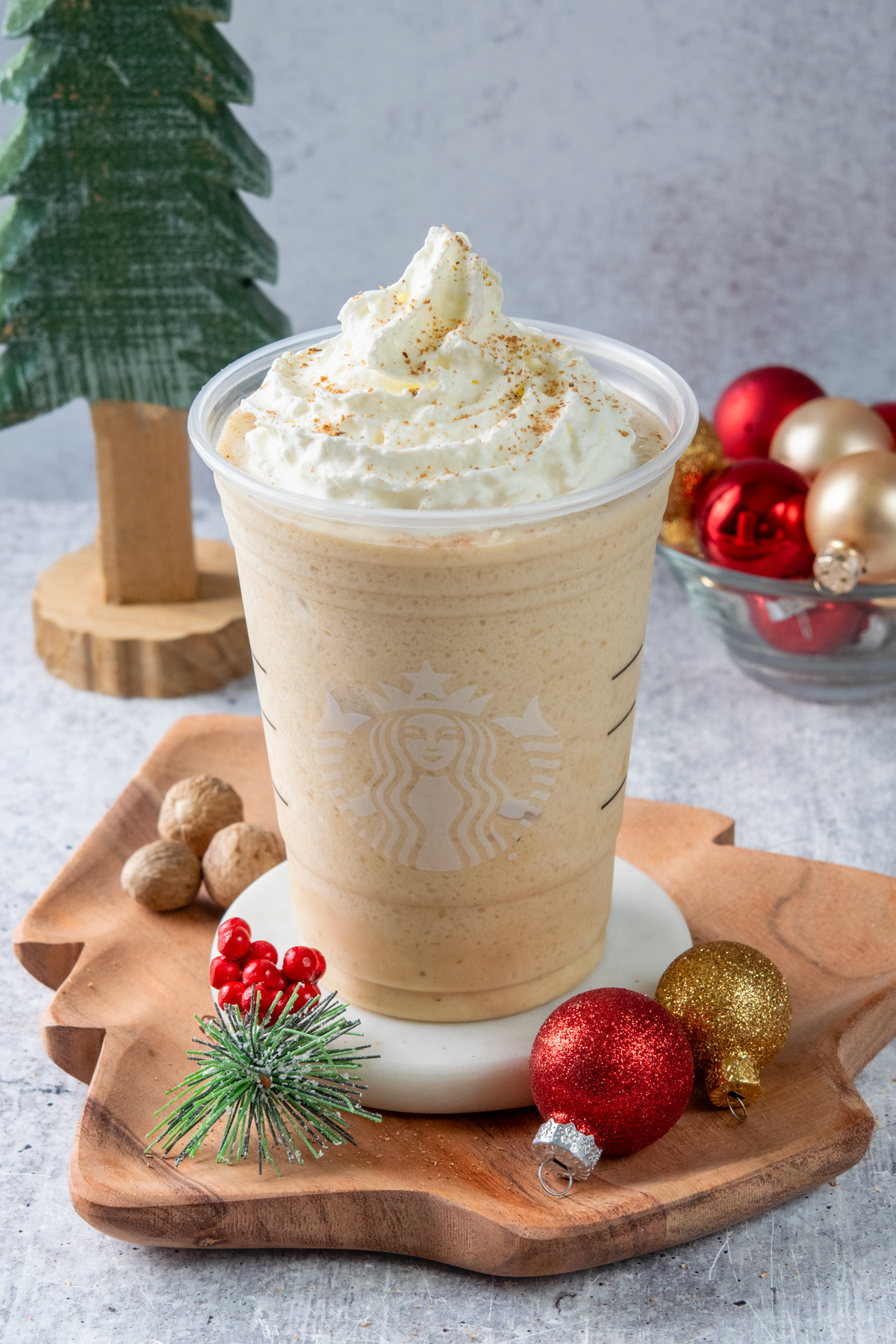 A thick and creamy eggnog frappuccino made at home in a Starbucks cup. The festive frappuccino is topped with a big swirl of whipped cream which has nutmeg sprinkled on top. Behind the copycat drink are some Christmas decorations including a Christmas tree and some small red and gold ball ornaments.