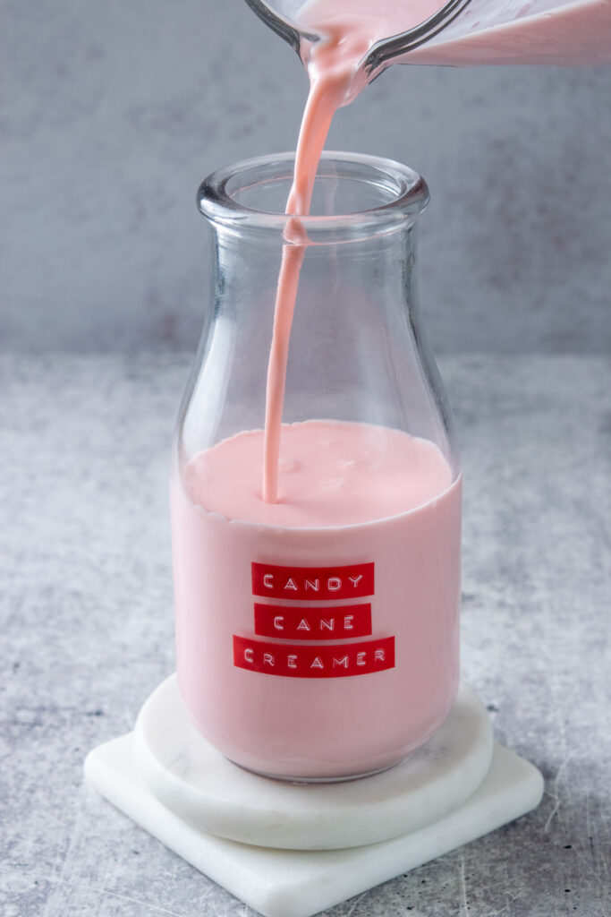 Pouring homemade candy cane syrup into a bottle that has a red label on it that says, "candy cane creamer."