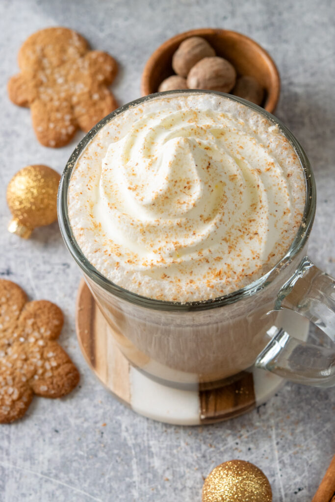 A homemade hot gingerbread chai latte topped with whipped cream and grated nutmeg. The cozy latte is being served with gingerbread cookies. The drink is surrounded by a few gold Christmas ball ornaments.