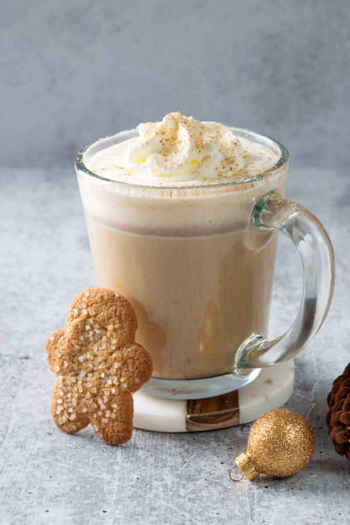Glass mug of hot gingerbread chai latte topped with whipped cream and nutmeg, with a gingerbread man cookie and gold Christmas ornament beside it.
