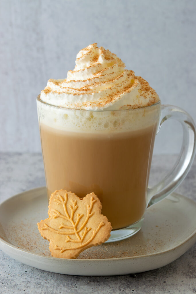 Homemade maple latte topped with whipped cream and cinnamon and one maple leaf shaped cookie next to it.