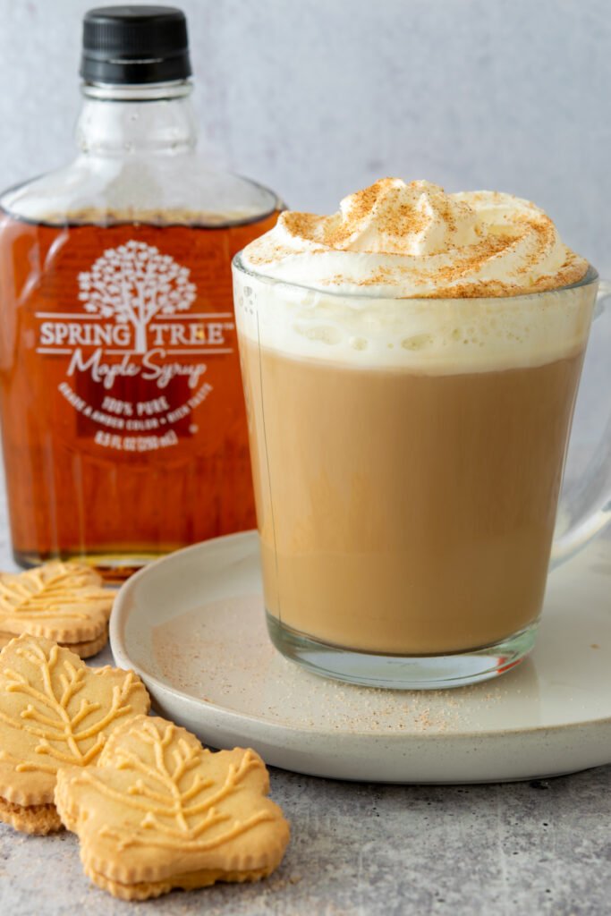 A bottle of pure maple syrup, maple leaf shaped cookies and a maple latte with whipped cream and cinnamon.