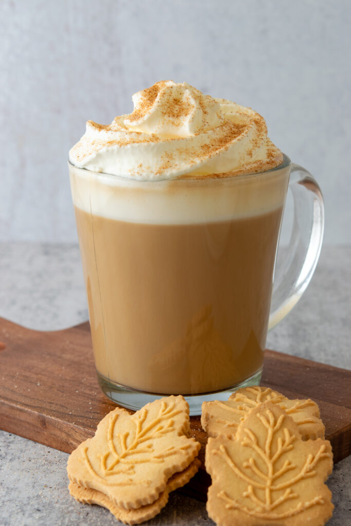 Maple latte topped with whipped cream and cinnamon. There are three maple leaf shaped cookies next to the hot latte.