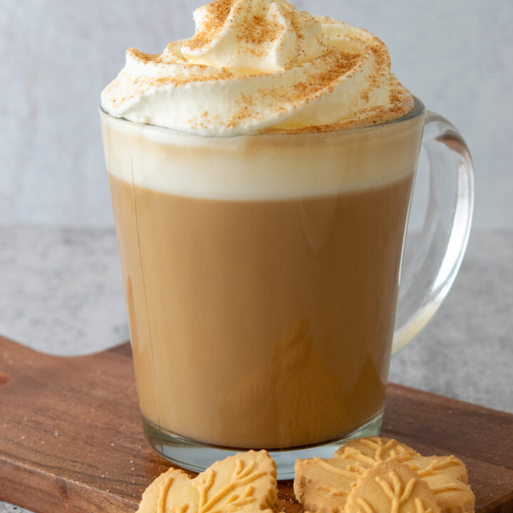 Maple latte topped with whipped cream and cinnamon. There are three maple leaf shaped cookies next to the hot latte.