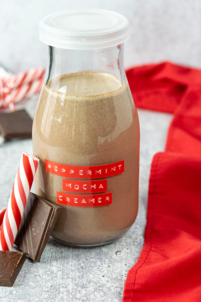 Bottle of homemade peppermint mocha creamer with a red label on it. There's chocolate and a peppermint stick leaning next to the creamer.