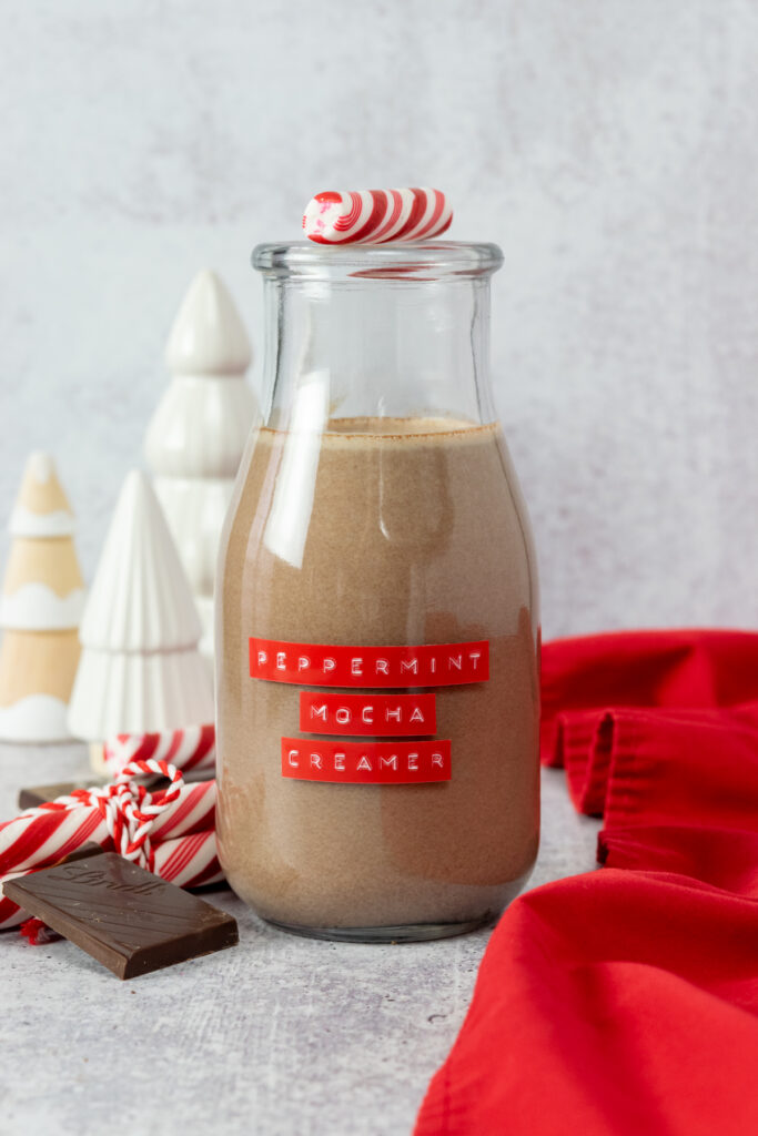 Homemade peppermint mocha creamer in a small glass milk bottle with a red label on it. There are some decorative Christmas trees, and some chocolate bars and peppermint sticks next to it.