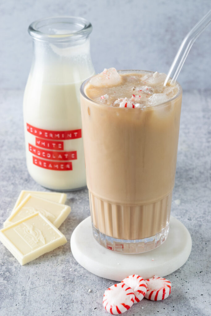 An iced peppermint mocha with pieces of crushed peppermint candy on top, sitting next to three starlight peppermint candies, three white chocolate Ghirardelli squares and a bottle of homemade peppermint white chocolate creamer.