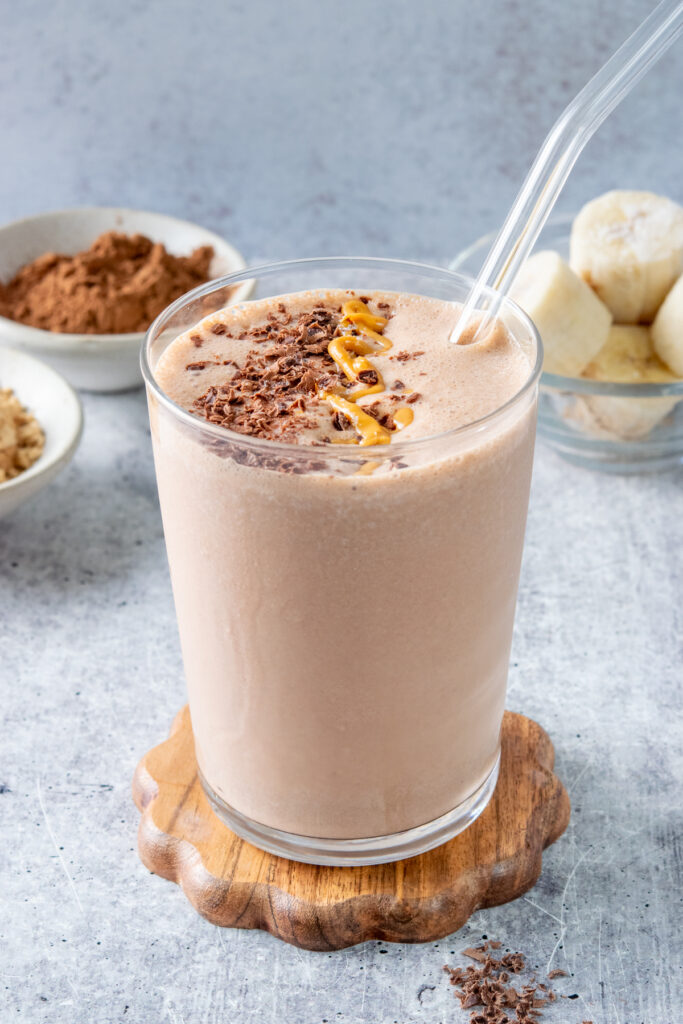 Rich and creamy chocolate peanut butter banana smoothie topped with chocolate and peanut butter, with bowls of powdered peanut butter, cocoa powder, and frozen banana pieces in the background.