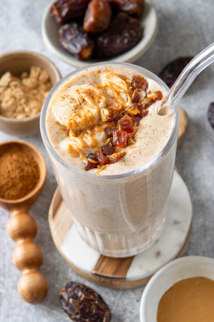 A banana date smoothie in a glass with a straw. The glass is on a coaster and surrounded by Medjool dates, a wooden scoop with cinnamon and a small bowl with peanut butter powder.