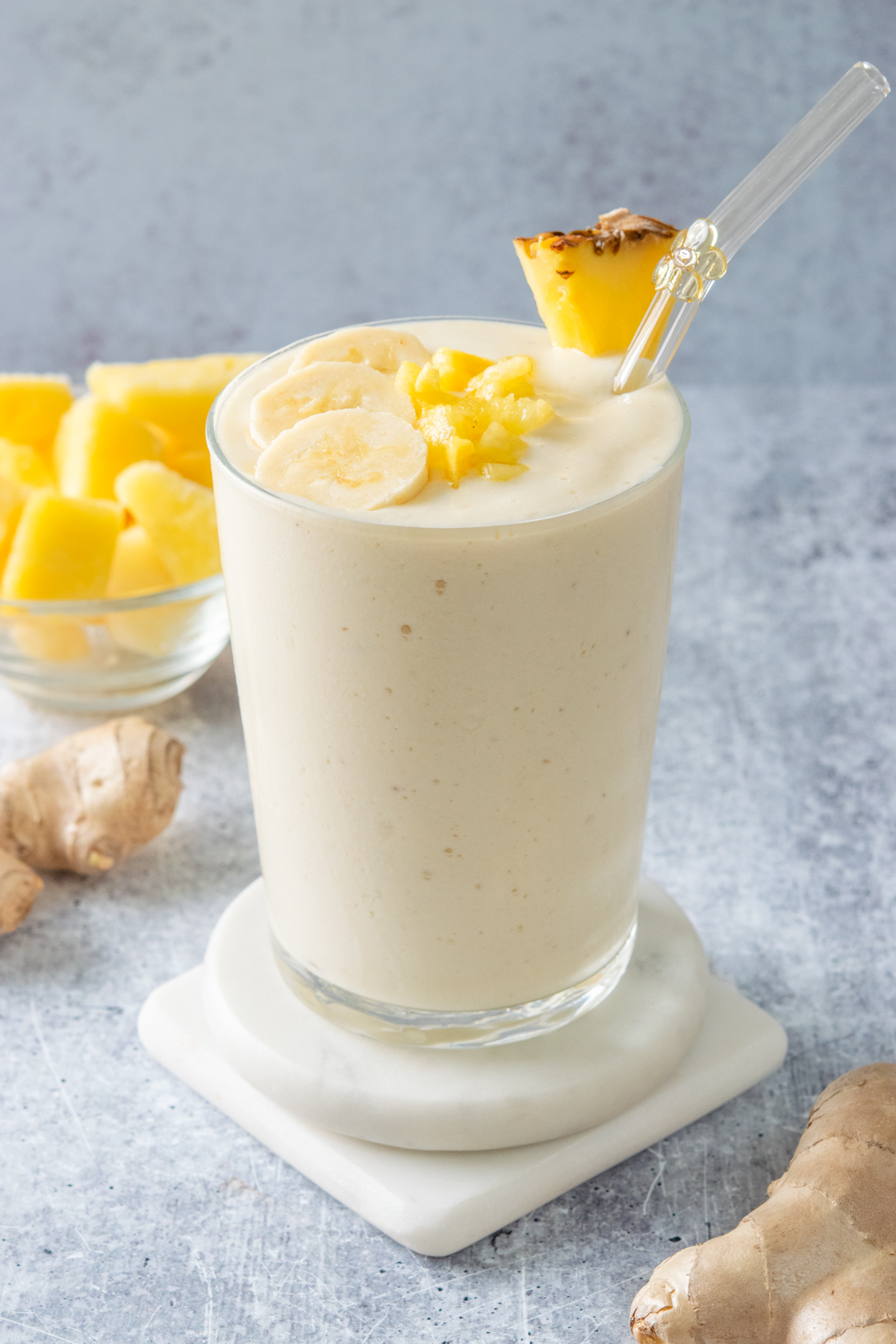 Creamy pineapple ginger smoothie in a glass with a glass straw, topped with banana slices and chopped pineapple, with fresh ginger root and pineapple chunks beside it.