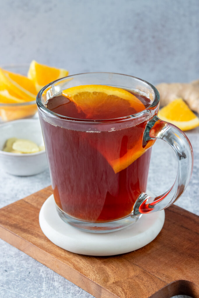 A cup of hot orange ginger black tea, near some sliced ginger root and an orange sliced into wedges.