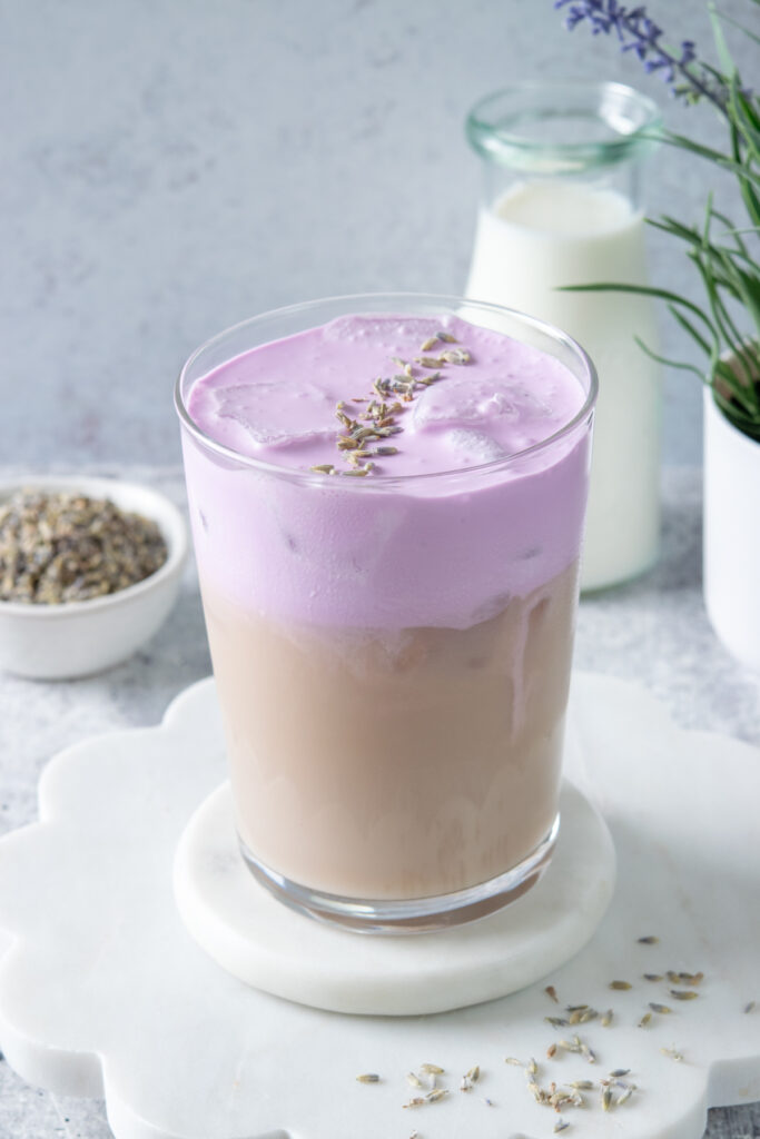 Layered iced lavender cream chai latte with light purple lavender cold foam over spiced chai, topped with dried lavender flowers, with milk pitcher and lavender plant in background.