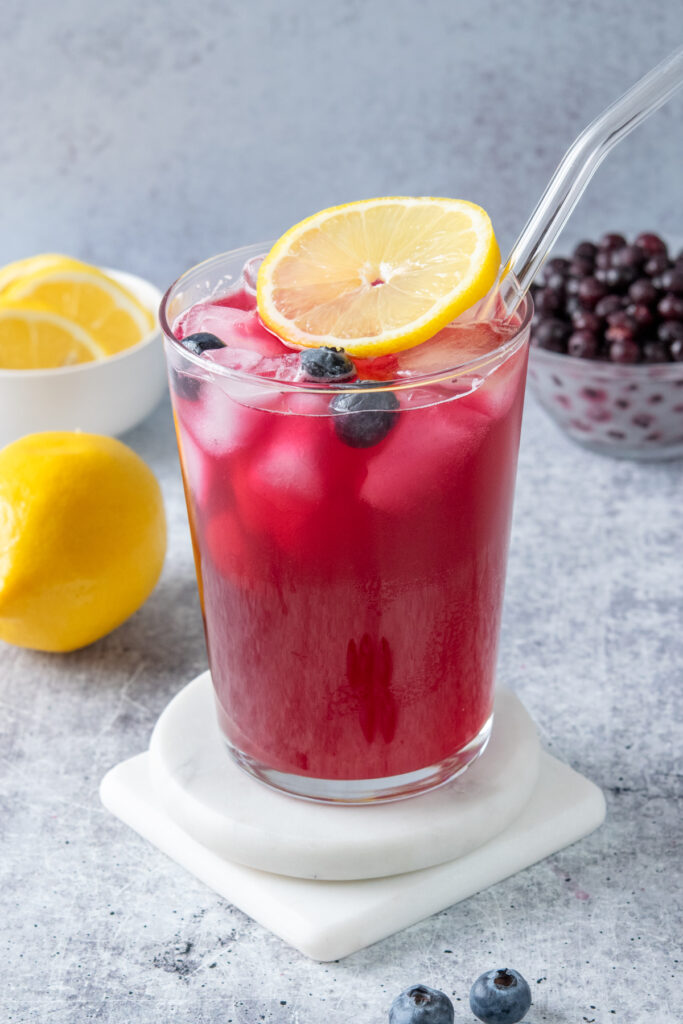 Glass of homemade blueberry lemonade garnished with fresh blueberries and a lemon slice with whole lemon and bowl of blueberries in the background.