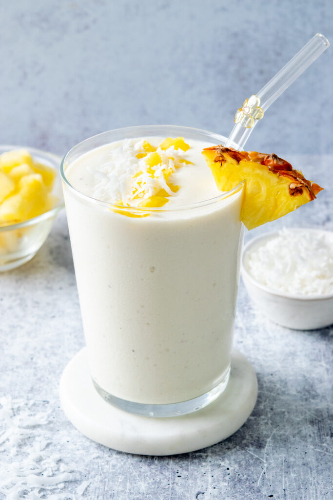 Creamy pina colada smoothie in a glass with a glass straw, garnished with a pineapple wedge and shredded coconut, with bowls of pineapple chunks and coconut in the background.