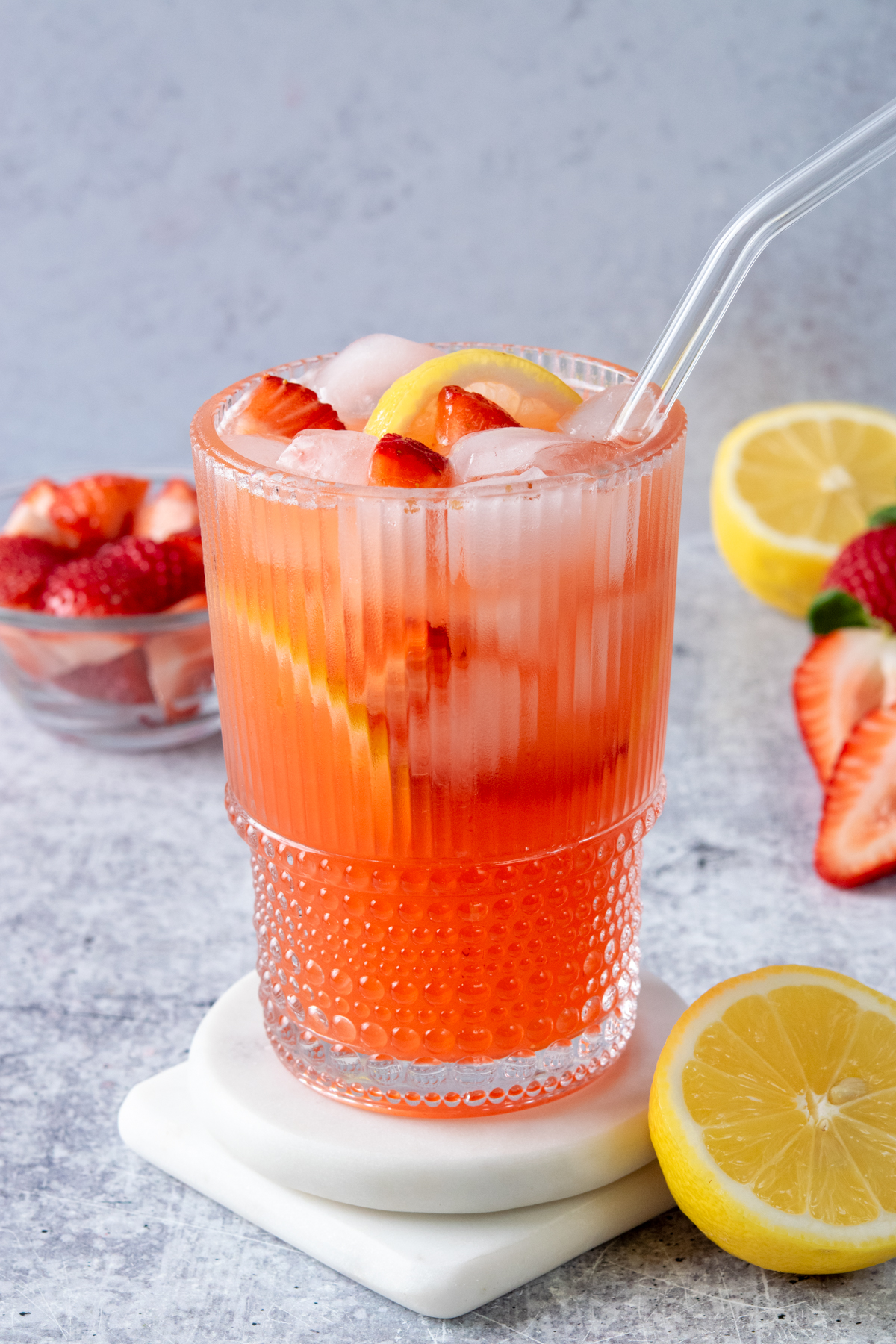 Ice-cold glass of homemade strawberry lemonade with fresh strawberry pieces and lemon slices, served beside a bowl of sliced strawberries and a halved lemon.
